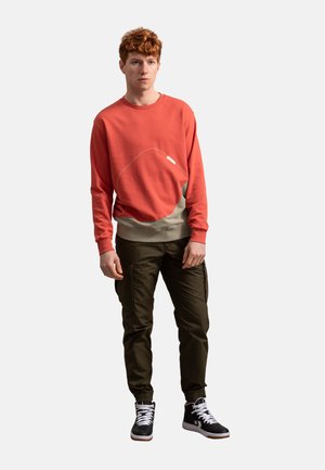 Felpa rossa con un design a linee bianche, pannello inferiore beige; abbinata a pantaloni cargo verde scuro e sneakers nere alte.