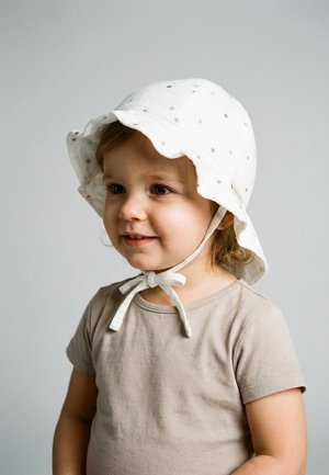 Toddler wearing a beige shirt and white sun hat with small dots, tied under the chin, smiling and looking slightly to the side.