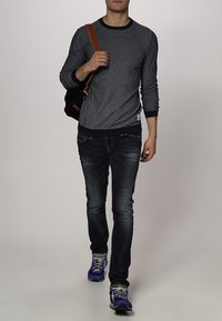 Grijze trui met ronde hals en een marineblauwe geribde zoom, gecombineerd met donkere jeans en paarse sneakers. Een zwarte schoudertas hangt over het lichaam.