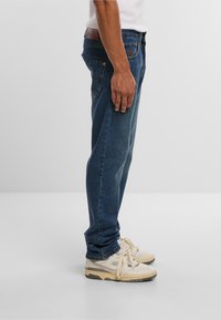 Blauwe denim jeans met een rechte snit, voorzien van koperen klinknagels en een bruine leren patch op de achterkant. Gepaard met witte sneakers.