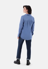Light blue long-sleeve shirt with a collar, buttoned cuffs, and a rounded hem. Styled with dark blue jeans and black ankle boots.
