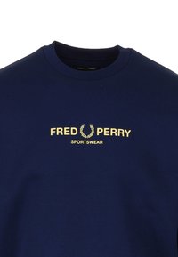 Felpa blu navy con scollo rotondo e polsini a costine. Presenta testo dorato: "FRED PERRY" con un logo di alloro e la scritta "SPORTSWEAR" sotto. Texture liscia.