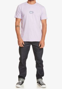 Light lavender cotton t-shirt featuring a small blue graphic design on the chest, paired with dark denim jeans and black sneakers.