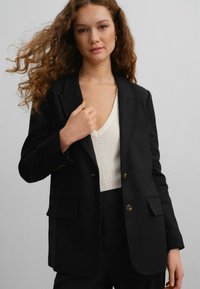Black tailored blazer with a fitted design, subtle texture, two front pockets, and gold buttons. Paired with a textured cream top.