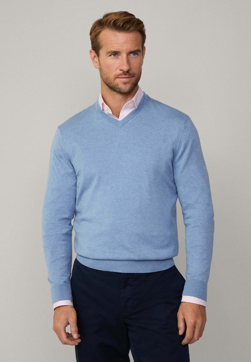 Hellblaues V-Neck-Sweatshirt aus weichem Material, mit gerippten Bündchen und Saum, getragen über einem hellrosa Kragenhemd. Marineblaue Hose vervollständigt den Look.