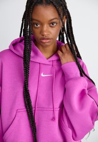 Sudadera rosa hecha de un tejido suave, que presenta un bolsillo frontal y cordones. Incluye un pequeño logo blanco de Nike en el pecho.