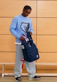 Tengerészkék hátizsák piros pántokkal és fehér betűkkel, több rekesszel és egy kör alakú logóval, egy kék pulóvert viselő személy tartja.