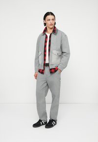 Jachetă din denim gri purtată peste o cămașă din flanel în carouri roșii și negre, asortată cu pantaloni gri și adidași negri cu dungi albe.