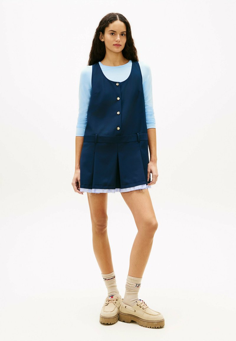 Femme aux longs cheveux foncés portant un haut à manches longues bleu clair sous une robe sans manches bleu marine boutonnée, des chaussettes beiges et des chaussures beiges épaisses.