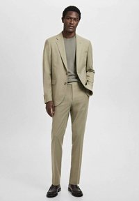 Ljusbeige kavaj och byxor över en grå skjorta. Slät tyg, skräddarsydd passform, en knapp på kavajen. Mörka skor med texturerad detalj.