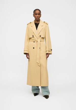 Donna che indossa un lungo trench beige con cintura sopra jeans larghi azzurri e scarpe nere con punta, in piedi contro uno sfondo neutro.