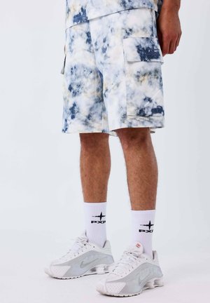 Uomo che indossa pantaloncini cargo tie-dye bianchi e blu, calzini bianchi con logo PXCP nero e scarpe da ginnastica Nike bianche con suole a molla.