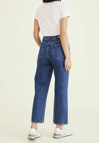 Vaqueros de mezclilla azules de talle alto con corte recto, cinco bolsillos, costuras visibles, combinados con una camiseta ligera a rayas y zapatillas blancas.