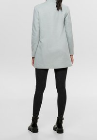 Abrigo gris claro con un diseño ajustado, mangas largas y un dobladillo ligeramente acampanado, que se lleva sobre unos jeans ajustados negros y botas de tobillo.