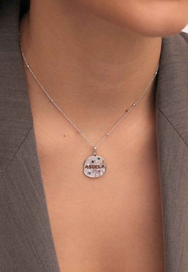 Collar de plata que presenta un colgante circular con la palabra "ABUELA" grabada. El colgante está decorado con piedras preciosas de colores y un acabado texturizado.