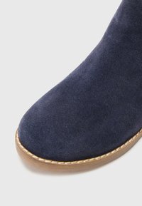 Bottine en suède bleu marine foncé avec un bout rond et une semelle extérieure contrastante en beige clair, ornée de coutures décoratives autour du bord.