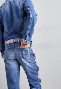 Chaqueta de mezclilla con mangas largas y un ribete difuso en contraste; jeans azules con un panel lateral púrpura distintivo y múltiples bolsillos.