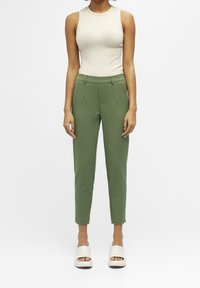 Débardeur beige côtelé associé à un pantalon vert ajusté et des sandales blanches à plateforme, mettant en valeur une coupe ajustée et des lignes épurées.