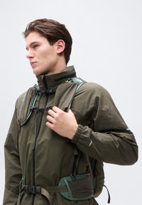 Νέος άντρας που φοράει πράσινο ελαφρύ μπουφάν και γιλέκο πεζοπορίας, προσαρμόζοντας το λουρί στον ώμο, κοιτάζοντας προς τα αριστερά σε απλό λευκό φόντο.