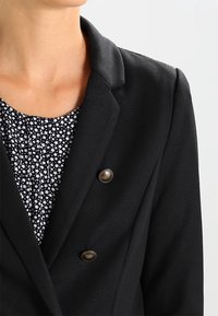 Svart blazer med slagkrage, dubbelknäppt design och mässingsknappar. Lags under en mönstrad blus med små vita stj ärnor.