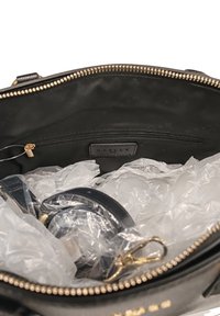Interno di una borsa in pelle nera con zip dorata. Contiene plastica spiegazzata e una tracolla arrotolata con un moschettone dorato. Etichetta visibile all'interno.