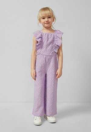 Młoda dziewczyna o blond włosach, ubrana w liliowy koronkowy kombinezon z falbanianymi rękawami i białe trampki, stojąca na tle jednolitego szarego tła.