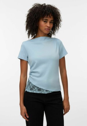 Femme aux cheveux bouclés portant un haut bleu clair à manches courtes superposé sur un tissu en dentelle, associée à un pantalon noir sur fond blanc.