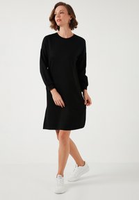 Black long-sleeve dress made of textured fabric, featuring a crew neckline and relaxed fit, paired with white sneakers on a light background.