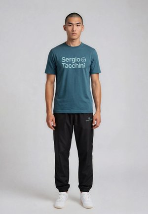 Joven de pie, vistiendo una camiseta verde azulado Sergio Tacchini, pantalones jogger negros con logo y zapatillas blancas, sobre un fondo claro y liso.