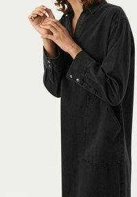 Czarna sukienka dżinsowa z kołnierzykiem, długimi rękawami i guzikami zatrzaskowymi. Posiada boczną kieszeń i luźny krój. Miękka tekstura i minimalistyczny design.