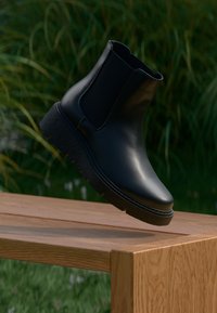 Schwarze Leder-Chelsea-Stiefel mit runder Zehenform, elastischen Seitenpaneelen und einer strukturierten Gummisohle, die auf einer Holzoberfläche platziert sind.