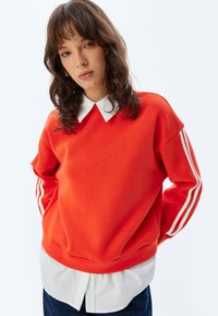 Sudadera roja con rayas blancas en las mangas, superpuesta sobre una camisa blanca de cuello. Presenta un corte holgado y un escote redondeado.