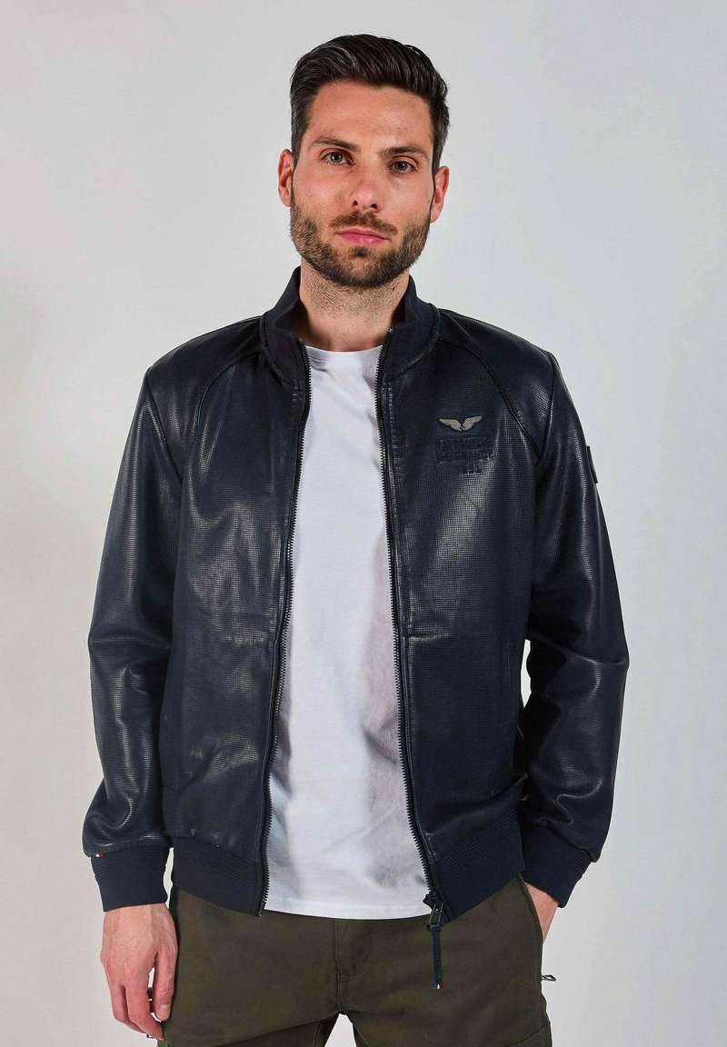 Veste bomber texturée bleu marine avec fermeture éclair, poignets côtelés et accent de logo sur la poitrine, associée à un t-shirt blanc et un pantalon olive.