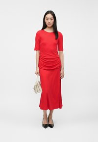 Κόκκινο φόρεμα με κοντά μανίκια, πτυχώσεις στη μέση, απαλή υφή, σε συνδυασμό με μια λευκή τσάντα και μαύρες γόβες με αιχμηρή μύτη.