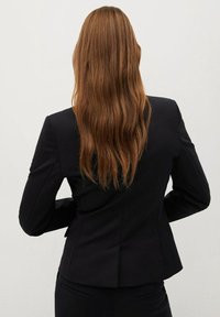 Blazer nero con una vestibilità su misura, caratterizzato da rever affilati, uno spacco posteriore e una texture liscia. La vista posteriore mostra capelli lunghi e ondulati.