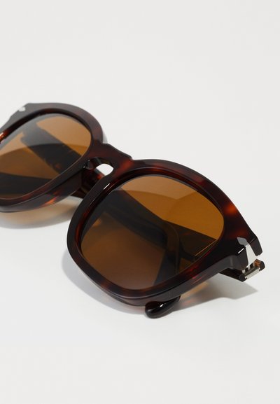 Tortoishell napszemüveg barna lencsékkel. Négyzet alakú, masszív keret, amely egyedi kivágásos dizájnnal és ezüst színű kiegészítőkkel rendelkezik.