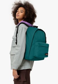 Zaino teal con tasca frontale, cerniera nera e etichetta di marca. Il modello indossa una giacca grigia chiara con dettagli viola.