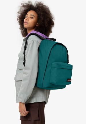 Türkise Rucksack mit einer Fronttasche, schwarzem Reißverschluss und einem Markenetikett. Das Modell trägt eine hellgraue Jacke mit lila Akzenten.