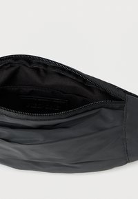 Μαύρο νάιλον τσαντάκι με φερμουάρ. Το εσωτερικό επενδύεται με λείο ύφασμα, διαθέτοντας μινιμαλιστικό σχεδιασμό και μία διακριτική ετικέτα με το λογότυπο.