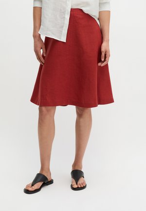 Frau, die einen knielangen roten Rock, ein weißes langärmeliges Hemd und schwarze offene flache Sandalen trägt, steht vor einem schlichten Hintergrund.