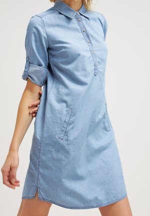 Kobieta ubrana w jasnoniebieską sukienkę z dżinsu z podwiniętymi rękawami, przednimi kieszeniami, zapięciem na guziki i małymi rozcięciami po bokach, pozująca z jedną ręką za sobą.