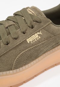 Sneaker in suede verde oliva con lacci piatti, marchio dorato e suola gommata traslucida, con dettagli di cucitura lungo il lato.