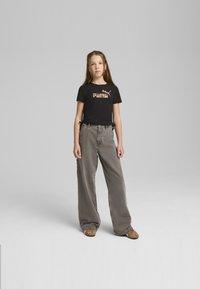 Camiseta negra con gráfico "PUMA", pantalones amplios grises y zapatos marrones. Diseño simple con lazos en los lados de la camiseta. Texturas suaves.