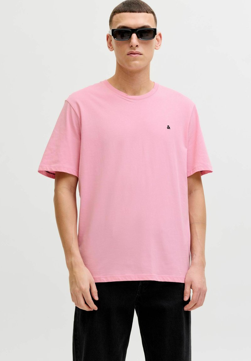Roze katoenen T-shirt met korte mouwen, ronde hals en een klein zwart logo op de borst. Gedragen met een zwarte broek en een zonnebril.