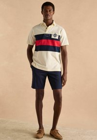 Polo cu nuanță crem, cu dungi orizontale albastre și roșii, având un patch cu logo; asortat cu pantaloni scurți bleumarin și pantofi de barcă bej.