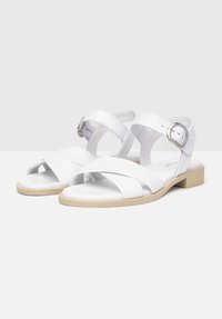 Des sandales blanches en cuir avec un design à lanières croisées, une sangle de cheville ajustable avec boucle, et une semelle beige légère. Style ouvert à l'avant.