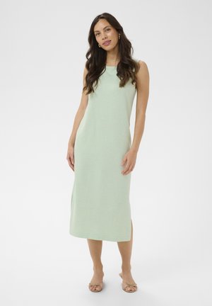 Kobieta o długich ciemnych włosach, ubrana w bezrękawnikową, jasnosiłozieloną sukienkę midi oraz beżowe sandały na obcasie, stojąca na białym tle.