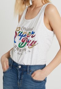 Camiseta sin mangas gris con el colorido logo de lentejuelas "Super Dry"; tejido suave, escote redondo y tirantes anchos; combinada con pantalones cortos de mezclilla azul.