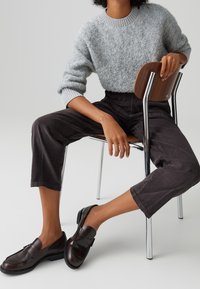 Personne portant un pull gris duveteux, un pantalon court foncé et des mocassins noirs assise sur une chaise en bois avec des pieds en métal sur un fond uni.
