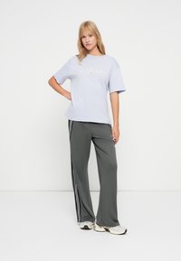 Blonde vrouw die staat, gekleed in een lichtpaarse Jordan t-shirt, donkergrijze trainingsbroek met witte strepen, en witte sneakers op een witte achtergrond.
